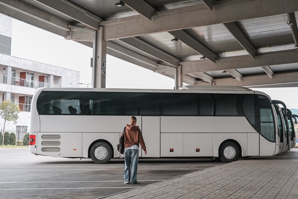 Frau mit Rucksack steht auf einem überdachten Parkplatz und schaut auf einen geparkten Reisebus. Im Hintergrund sind weitere Busse und ein modernes Gebäude zu sehen.