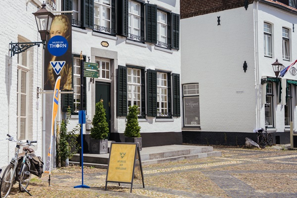 Fassade des VVV Thorn, der sich in einem weißen historischen Gebäude mit grünen Fensterläden befindet. Vor dem Eingang befinden sich eine Informationstafel und eine VVV-Fahne. Links von der Tür hängen ein blaues VVV-Schild und ein Banner des Museums Thorn. Auf der gepflasterten Straße lehnt ein Fahrrad an der Wand.